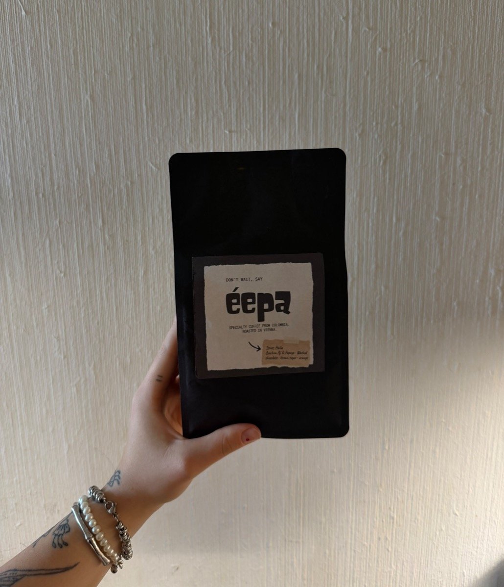 A hand holding up a black éepa coffee bag against a white textured wall