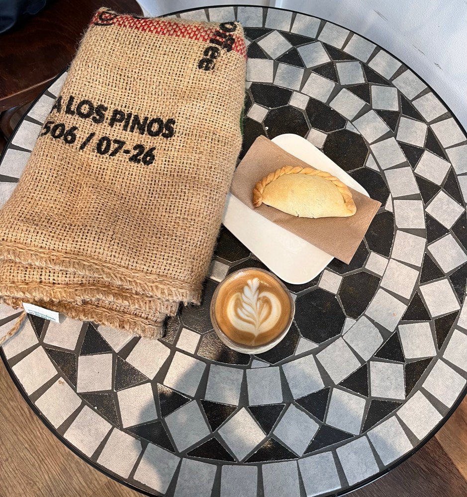 Jute sack printed Los Pinos next to a cortado and an empanada on a mosaic café table